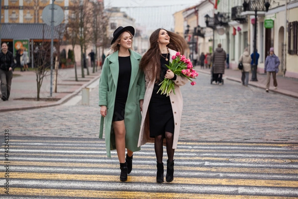 Obraz Two young Caucasian women walk together on a city street. One wears a green coat and the other a pink coat, both smiling and enjoying their time. One holds a bouquet of flowers