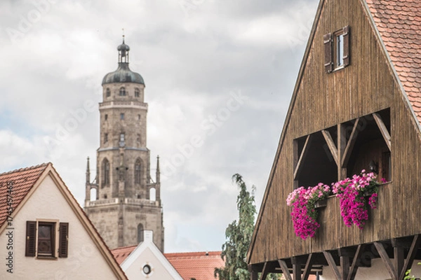 Fototapeta Historisches Haus mit Veranda und hängenden Balkonblumen an Giebelseite aus Holz vor unscharfem Kirchturm Daniel in historischer Altstadt in Nördlingen vor bewölktem Himmel.