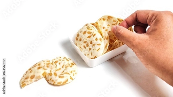 Fototapeta Hand taking Keripik Tempe or Tempeh Chips in white bowl. Isolated on white background with copy space. Traditional Indonesian food are made from soybeans mixed with rice flour then sliced ​​thinly.
