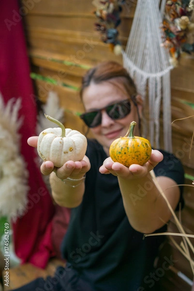 Obraz  smiling woman holding two small white baby boo and spotted munchkin pumpkins in an outdoor rustic fall display and looking at the camera, concept of seasonal marketing, diy craft, harvest festival