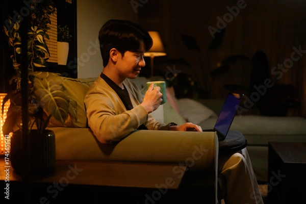 Fototapeta Relaxed remote worker sitting on sofa, smiling while using laptop and drinking coffee in dimly lit living room
