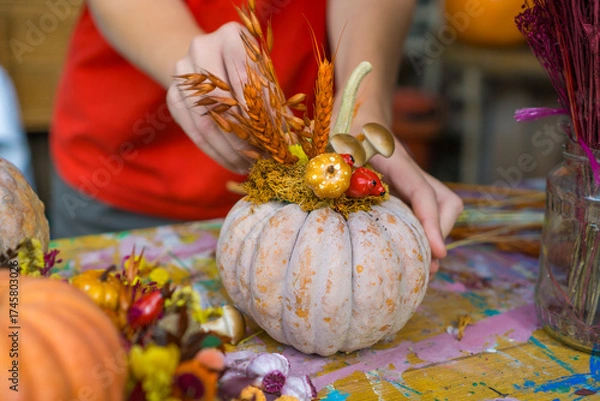 Obraz hands decorating a pale heirloom pumpkin with dried wheat stalks and ceramic mushrooms at a rustic workshop table, concept of diy craft, fall workshop, halloween, thanksgiving decor