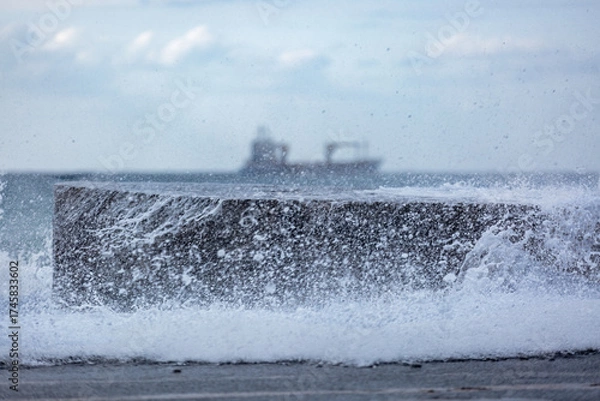 Fototapeta Waves crash on the stlny wall, in the background sea and ship in soft focus