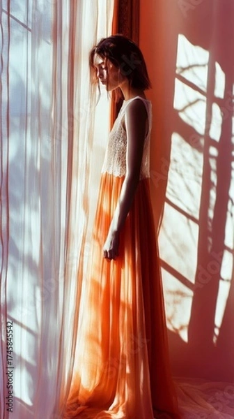 Fototapeta A woman in a long orange dress is standing in front of a window. The image has a warm and inviting mood, with the orange dress