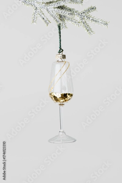 Fototapeta Minimal creative view of Glass of white wine Christmas decoration hang on snow green fir branch. New Year glass toy as wineglass with gold glitters. Festive minimal concept xmas party, winter holiday.