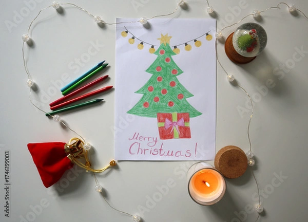 Fototapeta Flat lay of hand drawn Christmas tree with gift and Merry Christmas text, surrounded by crayons, candle, snow globe, and fairy lights. Childrens Christmas card. Festive content, kids projects, cards.