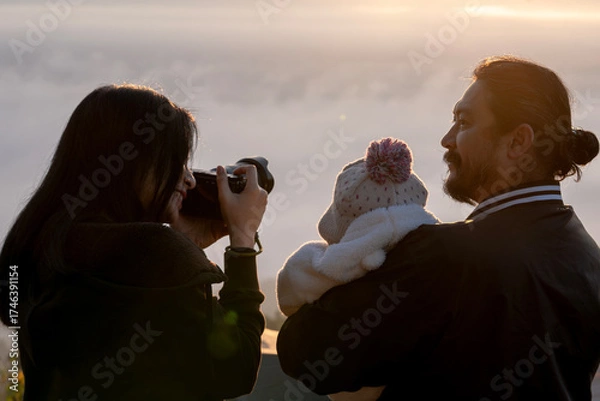 Fototapeta Parents and their little child pose for a photo on a misty mountain peak, enjoying warm sunlight in the winter season.