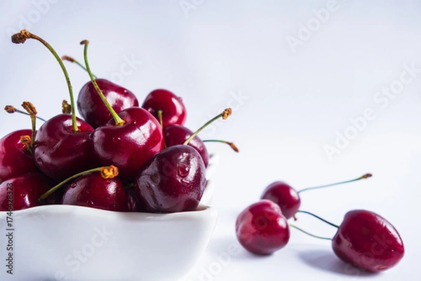 Fototapeta bright red cherries fill small white bowl with few cherries lying nearby on clean white surface, depicting freshness and summers bounty. scene looks simple and inviting. close up.