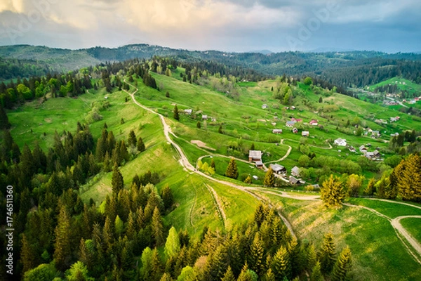 Fototapeta Aerial photograph of serene rural landscape in Ukraine, featuring rolling green hills, winding dirt roads, and scattered houses. Dense forests surround area, with dramatic, cloudy sky overhead.