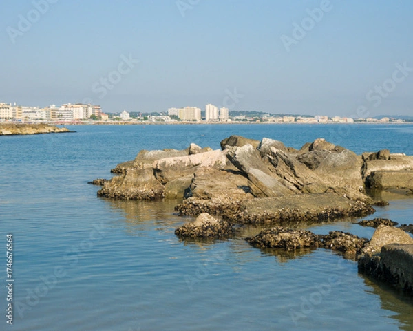 Obraz Low-angle view of a rugged, man-made rock breakwater in the calm sea. The modern skyline of the Italian resort town of Cattolica is visible in the background under a clear blue sky.