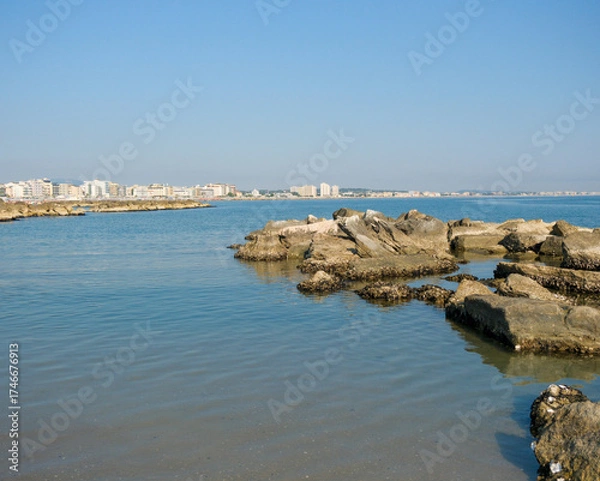 Obraz Low-angle view of a rugged, man-made rock breakwater in the calm sea. The modern skyline of the Italian resort town of Cattolica is visible in the background under a clear blue sky.