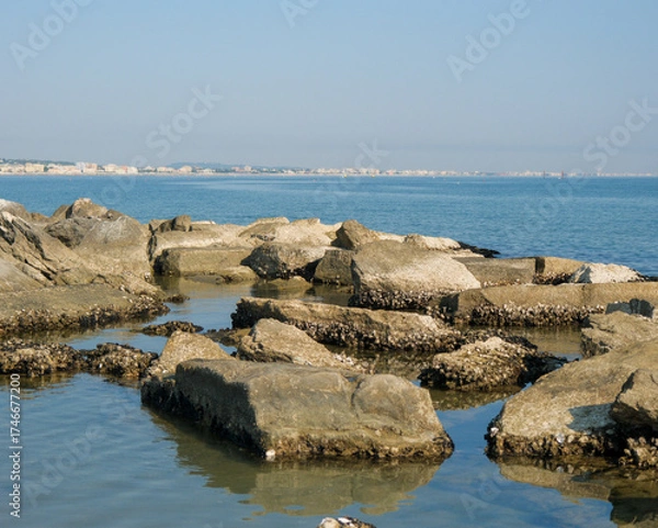 Obraz Low-angle view of a rugged, man-made rock breakwater in the calm sea. The modern skyline of the Italian resort town of Cattolica is visible in the background under a clear blue sky.
