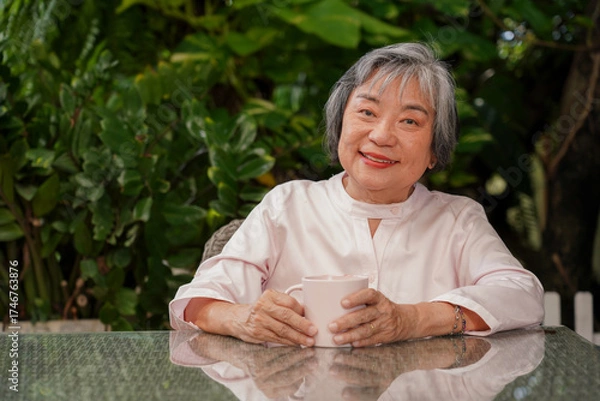 Obraz Portrait of a happy senior Asian woman smiling while holding a cup of coffee or tea in the garden. Enjoying a relaxed and healthy retirement lifestyle outdoors.