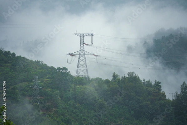 Fototapeta Power lines stand tall on green hillsides of Yichang, China, disappearing into the misty clouds, a landscape of energy infrastructure.