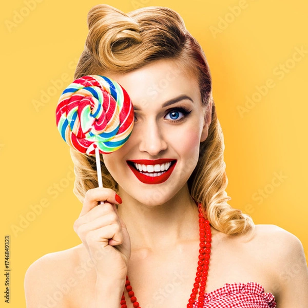Fototapeta Half face portrait of happy expressive smile woman holds lollipop in front of one eye. Cute pin up girl in retro style image. Isolated yellow background. Ophthalmology concept. Square