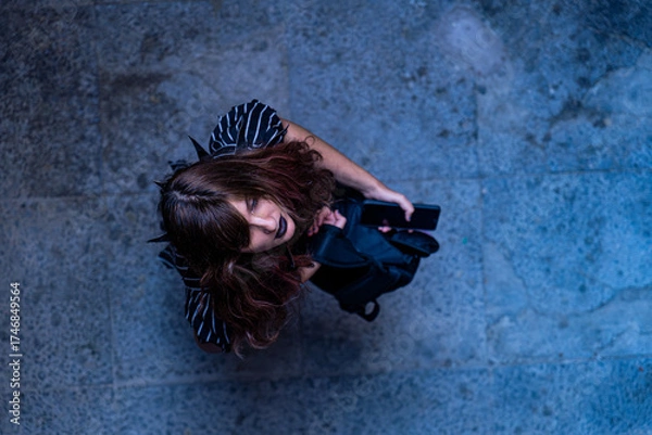 Fototapeta Top view of a young woman in dark clothes holding a phone and backpack, standing on a textured blue pavement, eyes closed, evoking nostalgia and melancholy.