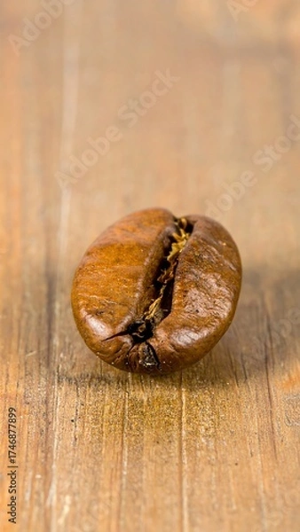 Obraz A close-up shot features a single, roasted coffee bean resting atop a textured, light-brown wooden surface. The bean?s dark-brown exterior reveals a slight crack