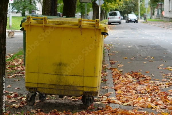 Fototapeta Large Yellow Waste Bin on Urban Street Curb with Autumn Leaves
