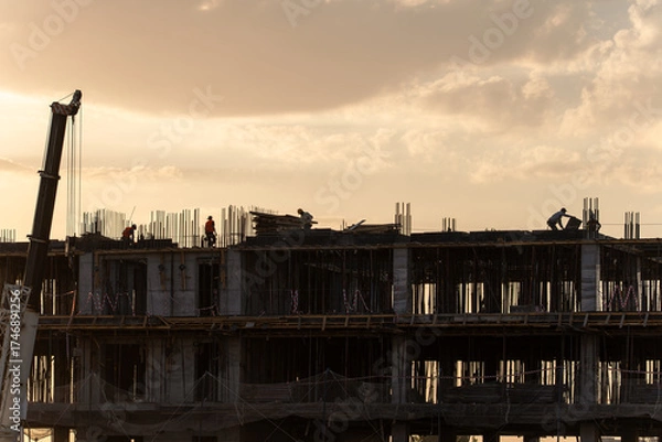 Fototapeta Silhouettes of construction workers on a building under construction against a beautiful, cloudy sky during the golden hour. A crane is visible on the left side of the frame.