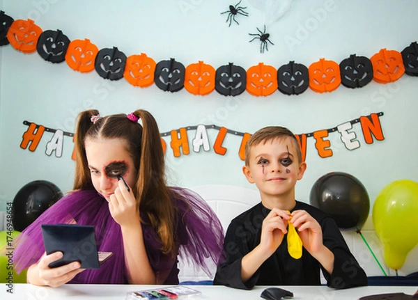 Fototapeta Two kids getting ready for Halloween. One girl applies makeup while boy inflates balloon.