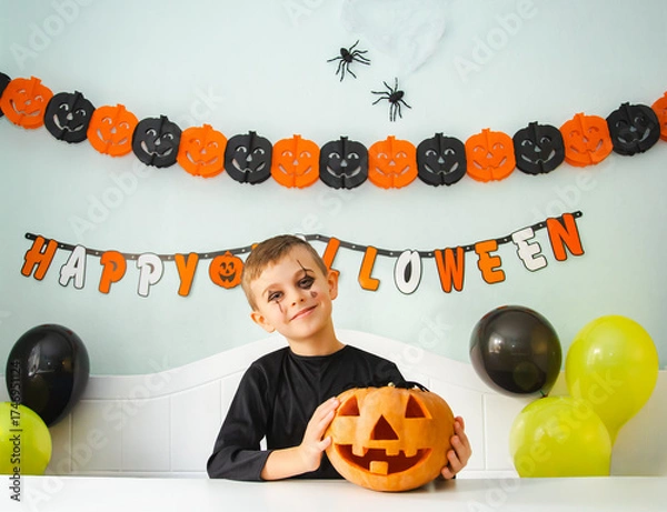Fototapeta Young boy in black costume holding carved jack-o’-lantern at Halloween party.