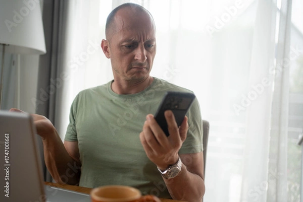 Fototapeta Shocked man looking at smartphone screen while using laptop working from home