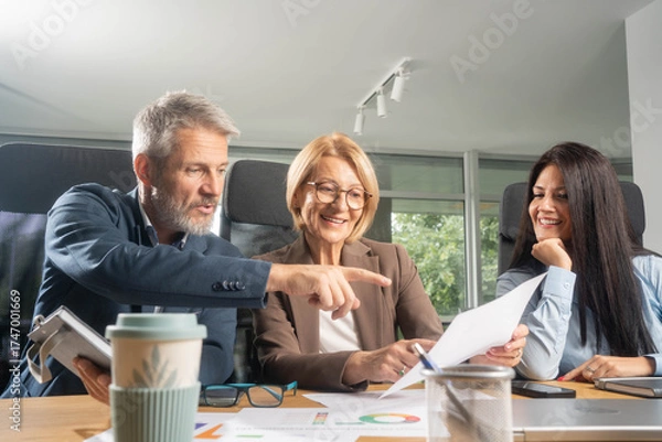 Fototapeta Three busy happy middle aged professional business man and two women executive leaders team using laptop working on computer at work desk having conversation on financial project at meeting in office.