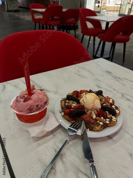 Fototapeta Plate with waffle, ice cream, strawberries, and cookies next to a cup of strawberry ice cream on a marble table with red chairs.
