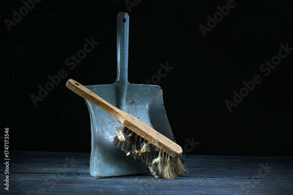 Fototapeta Old hand brush with wooden handle and a lot of dirt leaning on an aged metal dustpan against a dark background, still life of vintage cleaning tools, copy space
