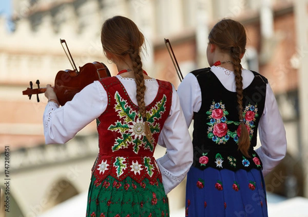 Fototapeta Two young girls in traditional folklore costiums embroidered with unique traditional pattern stand on their back playing violins