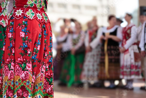 Fototapeta Woman in traditional folklore embroidered costume seen partianlly on stage, in the background group of people in traditional folklore clothes in soft focus