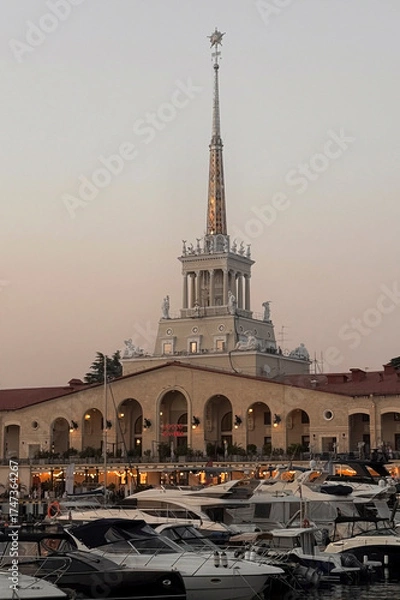Fototapeta A grand tower stands above a bustling marina filled with yachts near sunset in Sochi