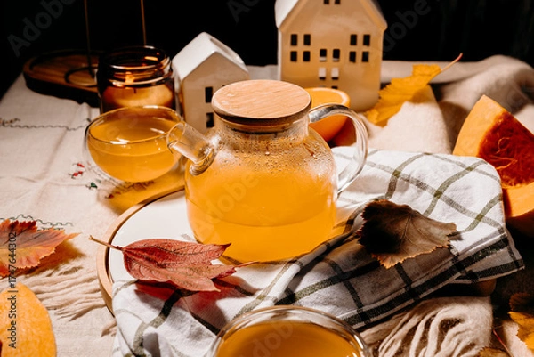 Fototapeta Transparent teapot filled with bright orange pumpkin tea surrounded by candles and autumn leaves. Cozy fall still life with warm steam.