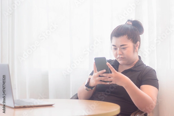 Obraz Young woman using smartphone while sitting at a table next to a laptop in a bright, airy room during the day
