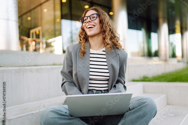 Obraz Portrait of a stylish woman with a laptop sitting on the steps in front of a modern office building. A beautiful woman in glasses works remotely outdoors. Freelancing concept, weekend.
