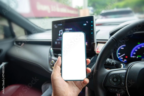 Fototapeta A person holding a smartphone with a blank white screen inside a modern car, focusing on the steering wheel and dashboard. Concept for car navigation app, mobile technology, safe driving awareness.