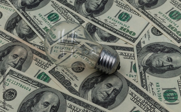 Fototapeta A light bulb against a background of hundred-dollar bills. A concept for saving on electricity. 