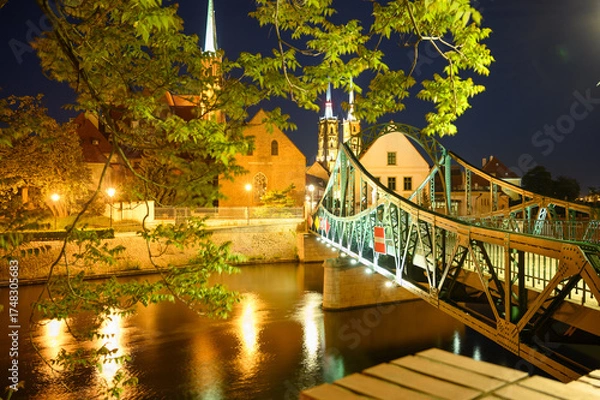 Fototapeta Night scene of the illuminated Tumski Bridge (Most) and Cathedral Island (Ostrów Tumski) in Wroclaw. Poland. Street lights and reflections in the Odra River create a romantic and peaceful atmosphere.