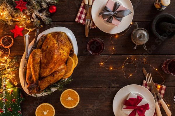 Fototapeta Romantic New Year dinner for two persons. Baked chicken as the main holiday dish on a table decorated with garland. Cone, dried oranges, plates, knife, fork furry spruce and two gifts on a table.