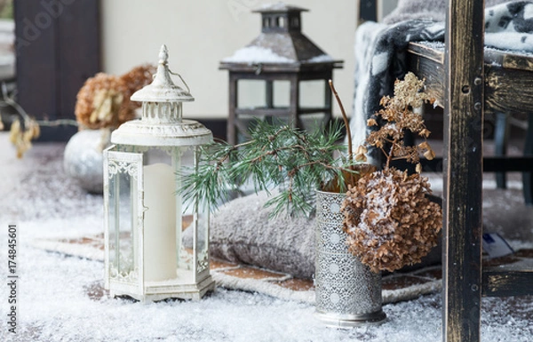 Fototapeta Outdoor new year decoration white lamp with the candle near wooden bench with artificial snow. Gray pillows and vases with plant on floor. Warm cozy atmosphere of holiday.