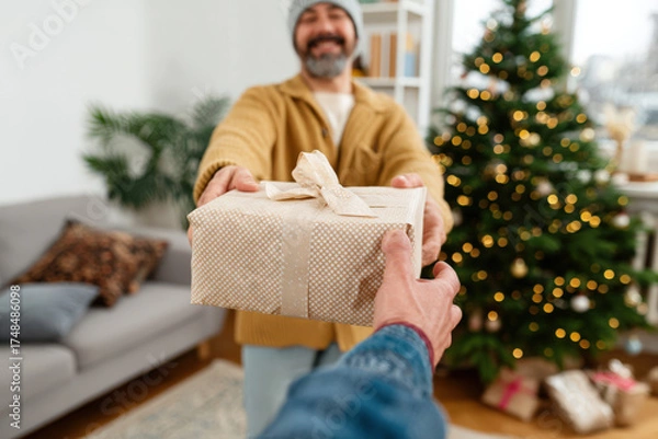 Fototapeta Smiling man in cozy sweater joyfully handing a beautifully wrapped gift to another person in a festive living room decorated for the holiday season with a Christmas tree