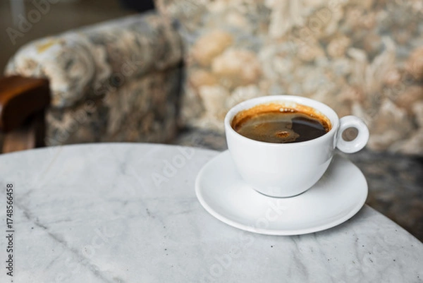 Fototapeta A white cup of black coffee americano in a cozy atmosphere of a cafe with copy space
