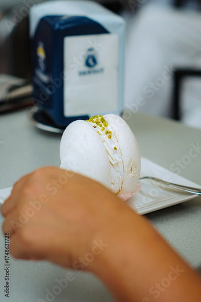 Fototapeta Pistachio meringue on white plate, tempting indulgence. Close-up reveals airy texture and vibrant green filling. A hand reaches for a fork, suggesting enjoyment. Subtle branded details in background.