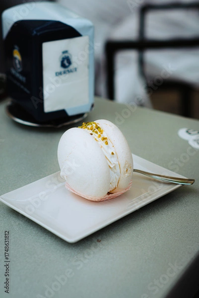 Fototapeta Pistachio macaron on a white plate, showcasing creamy filling and soft pink hues. Silver spoon included. Blurred cafe backdrop with architectural details evokes ambiance.