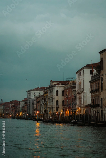 Obraz A picturesque view along a Venetian canal as dusk settles. Warm lights glow from the historic buildings, reflecting beautifully on the water's surface creating a magical and tranquil atmosphere.
