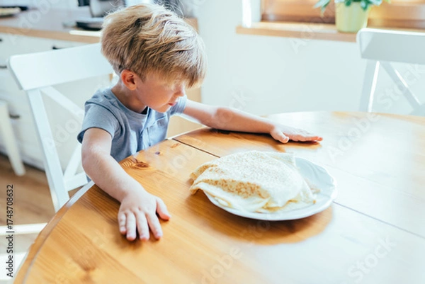 Fototapeta Little boy sitting at kitchen table waiting for pancakes on plate