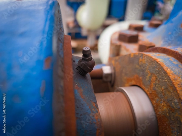 Obraz Close-up of a grease fitting (zerk) on rotating industrial machinery. Visible rust, bolts, and motion blur highlight active lubrication in a heavy-duty environment.