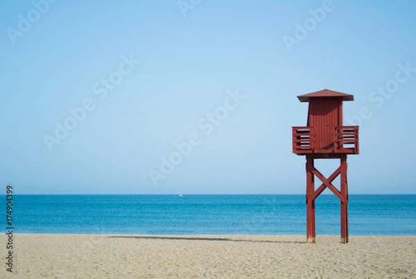 Fototapeta Red wooden lifeguard tower on the abandoned beach at Benalmadena, Malaga province, Spain. Beautiful view of the sea and sandy beach on sunny summer day with copy space.