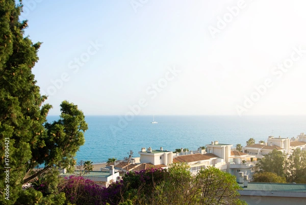 Fototapeta A view to Mediterrain sea from a view point over the tile roofs of white houses and hotels, green trees and pink violet colorful bougainvillea flowers at the foreground.