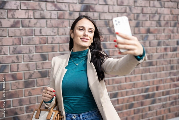 Obraz A smiling woman in a stylish outfit takes a selfie against a textured brick wall, showcasing happiness and confidence.